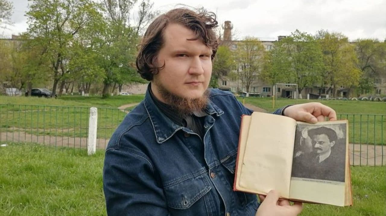 Bogdan Syrotiuk outdoors, holding a book open to a portrait of Leon Trotsky